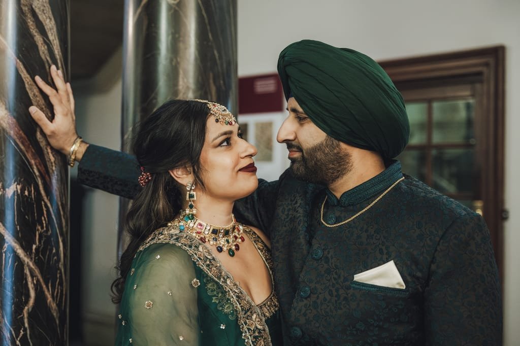 A couple dressed in traditional attire stands close together inside a building, smiling at each other. Captured by a Guildhall Bath wedding photographer, the man is wearing a turban and a dark outfit, while the woman is in a green embellished dress.