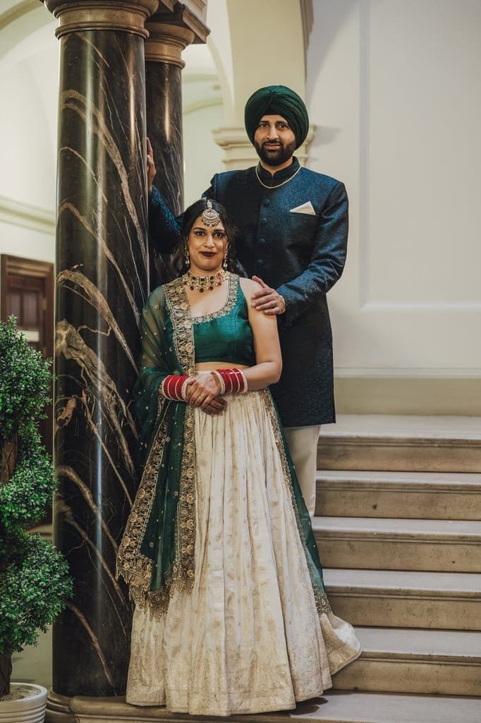 A couple in traditional Indian attire stands on a staircase, embodying wedding traditions from around the world. The woman dazzles in a green and white outfit while the man looks regal in a dark blue sherwani and green turban.