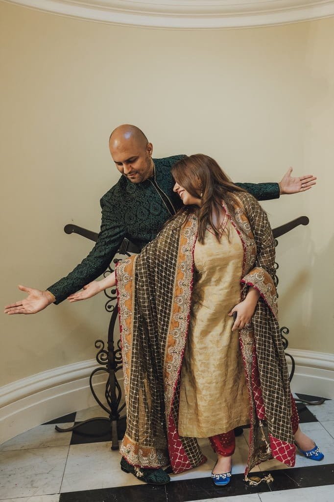 A man and woman pose playfully, extending their arms towards each other. Both wear traditional attire; he in a green outfit and she in a gold dress with a patterned shawl, embodying the rich essence of wedding traditions from around the world.