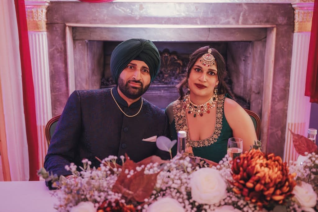 A couple in traditional attire sits at a decorated table with flowers and two bottles in front of a fireplace, embodying wedding traditions from around the world.
