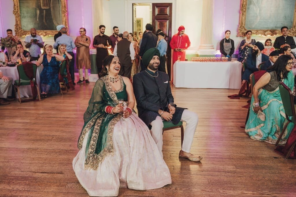 A couple dressed in traditional attire sit and laugh together at a wedding reception, embodying wedding traditions from around the world, with guests and servers bustling in the background.