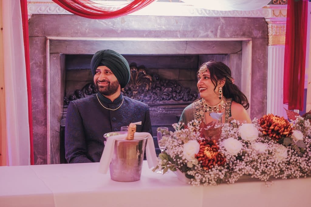 A man wearing a turban and a woman in traditional attire sit at a table adorned with floral arrangements and an ice bucket, smiling. Behind them, a decorated fireplace adds to the elegant ambiance, reflecting wedding traditions from around the world.