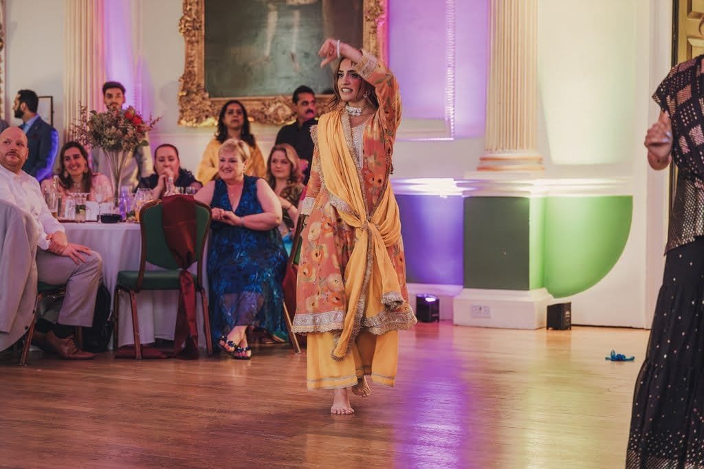 A woman dressed in traditional attire dances in a decorated hall, embodying wedding traditions from around the world, while seated guests watch her performance.