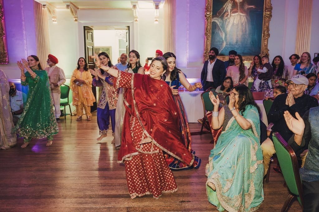 A group of people in colorful traditional attire are dancing and clapping in a decorated indoor setting, showcasing vibrant wedding traditions from around the world, with many onlookers enjoying the performance.