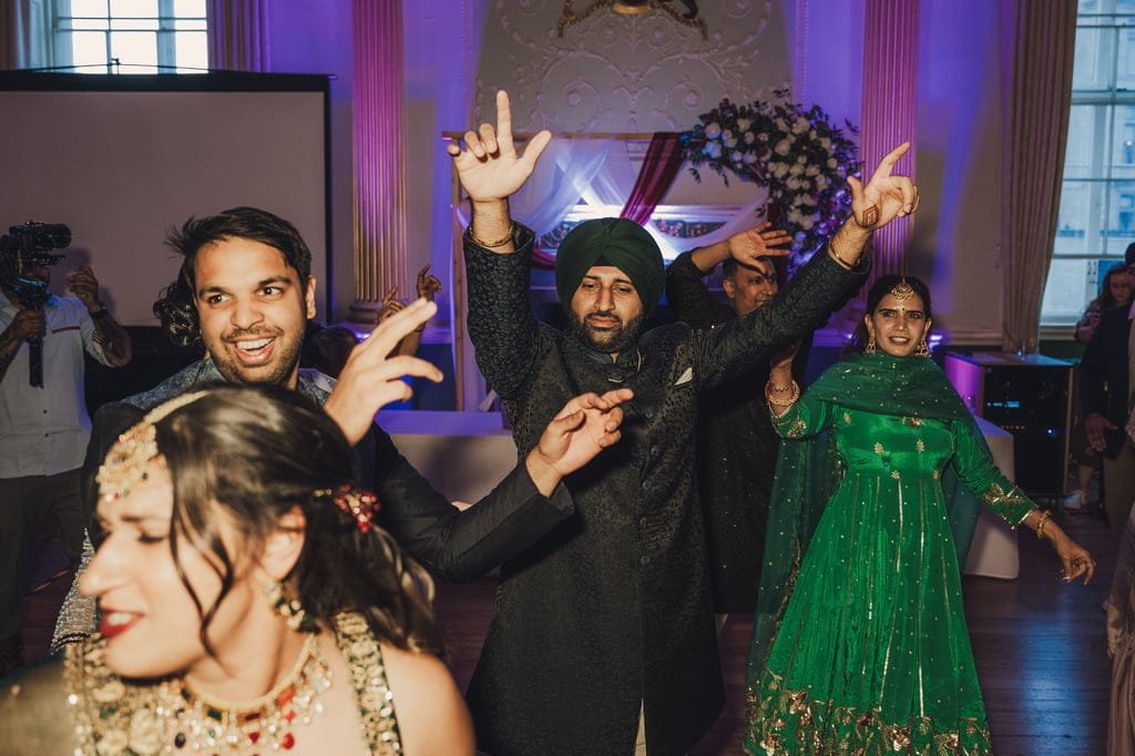 People are dancing and celebrating at an indoor event. Some are wearing traditional clothing, with one person in a green outfit. A photographer using Fuji Cameras for weddings captures the joyous moments as decorations and a flower arrangement adorn the background.