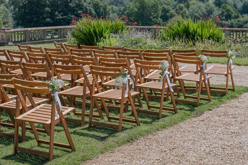 Rows of wooden chairs arranged outdoors on grass, adorned with white flowers and ribbons, capture the essence of an Elmhay Park wedding. The scenic garden and stone railing in the background create the perfect backdrop for stunning wedding photography.