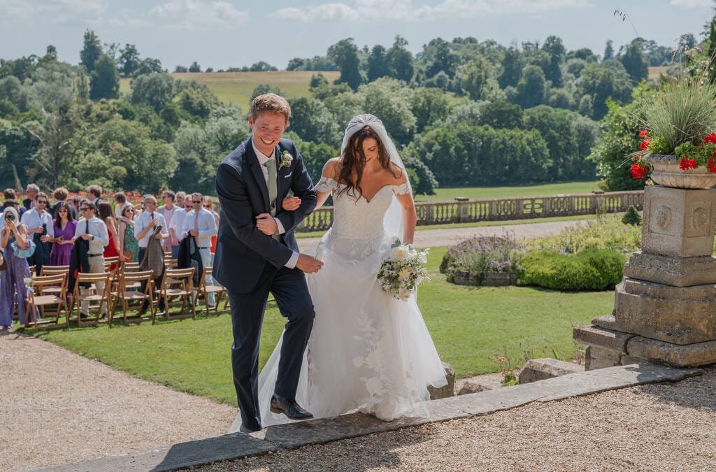 A bride and groom walk up stone steps outdoors at Orchardleigh House, holding hands and smiling, with trees and guests in the background.