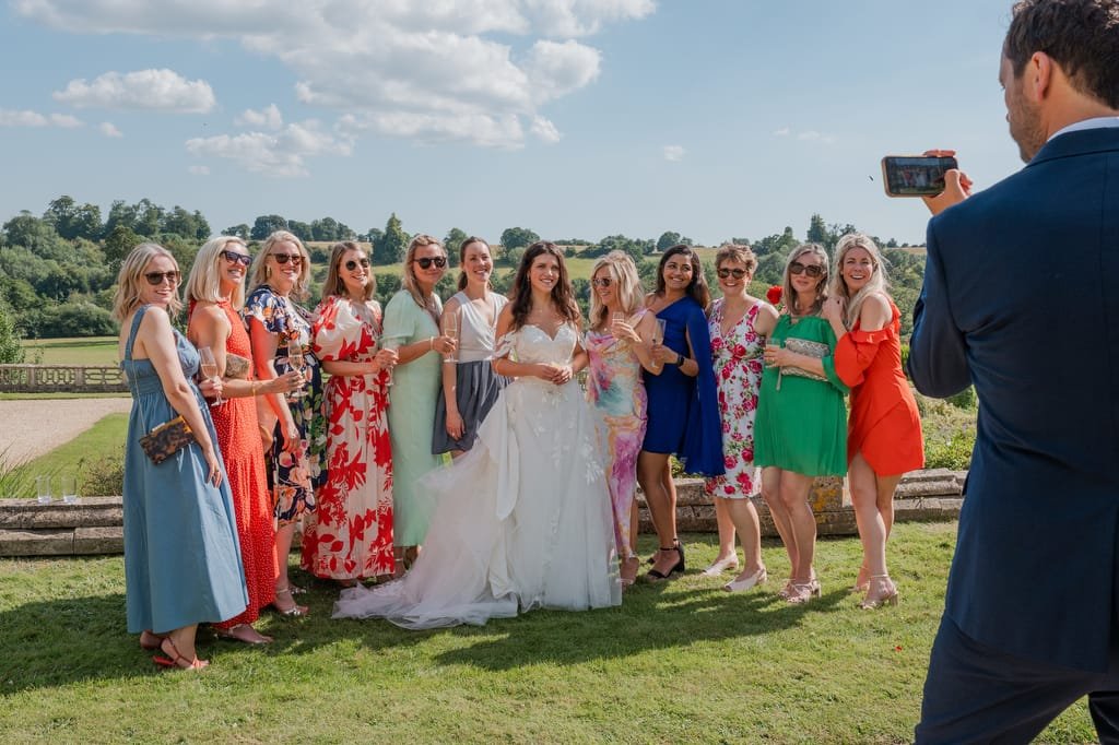 A group of women, one in a wedding dress, pose for a photo outdoors while a man captures the moment with his phone. The backdrop of Orchardleigh House features a scenic landscape with trees and a partly cloudy sky.