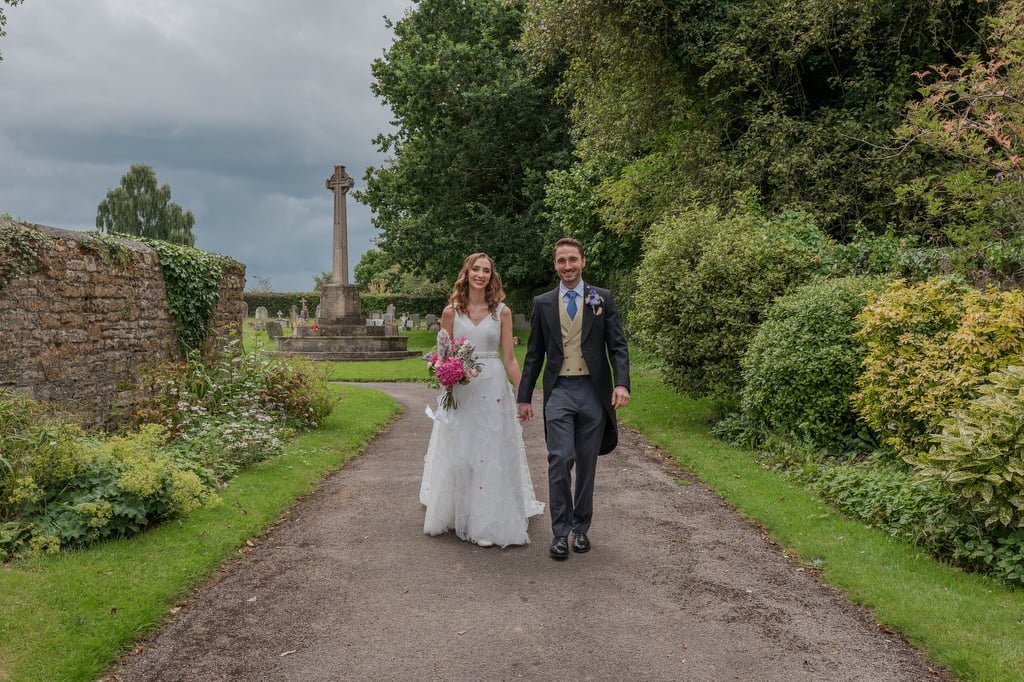 A bride and groom walk hand in hand down a tree-lined path at Pythouse Weddings, surrounded by stone walls and a large cross monument. The bride holds a bouquet of pink flowers.