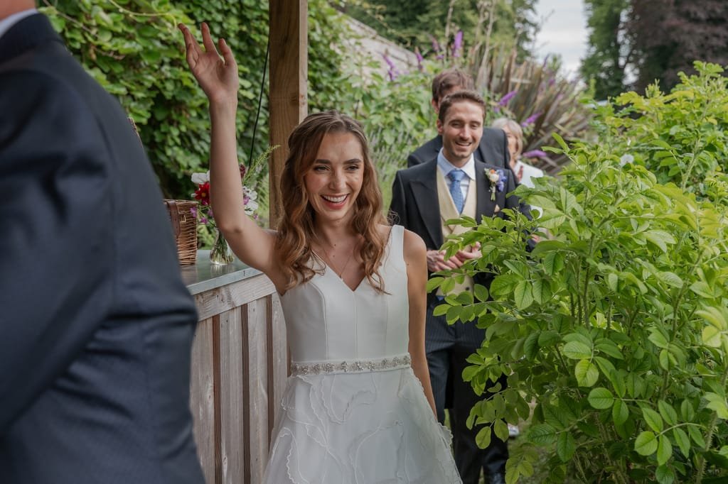 A bride in a white dress waves while smiling, with a groom in a suit standing behind her, surrounded by greenery at Pythouse Weddings.