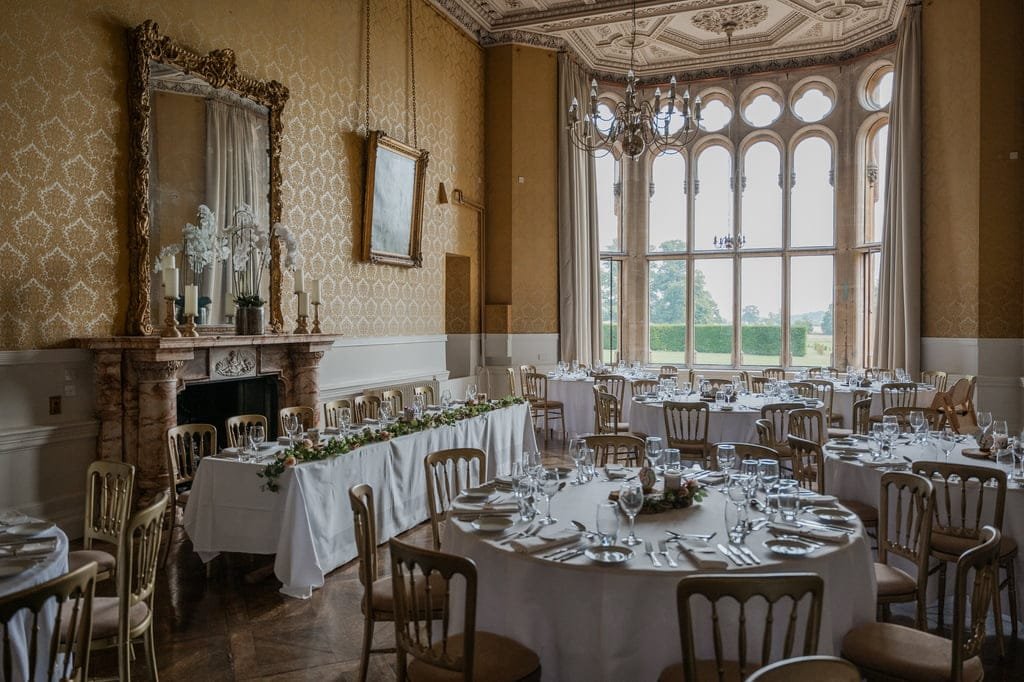 A formal dining room with ornate decor, round tables, white tablecloths, and wooden chairs set for a meal. Tall windows, a fireplace, and chandeliers complete the elegant setting, perfect for capturing by a Grittleton House photographer.