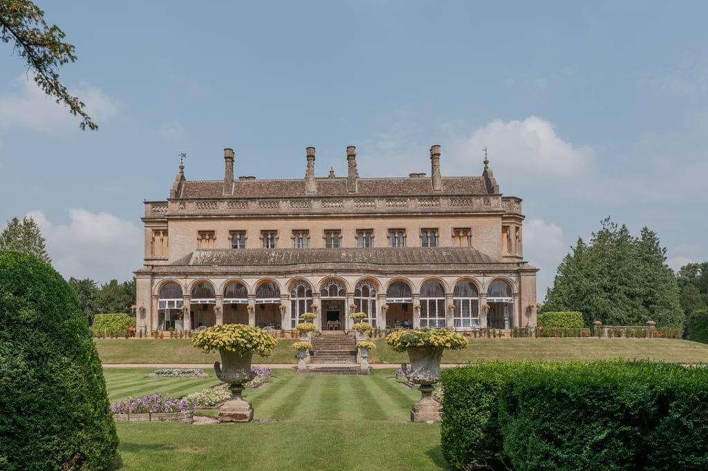 A large, historic building with an ornate façade and columns, Grittleton House is set in well-manicured gardens and surrounded by greenery, under a clear blue sky—truly a photographer's dream.