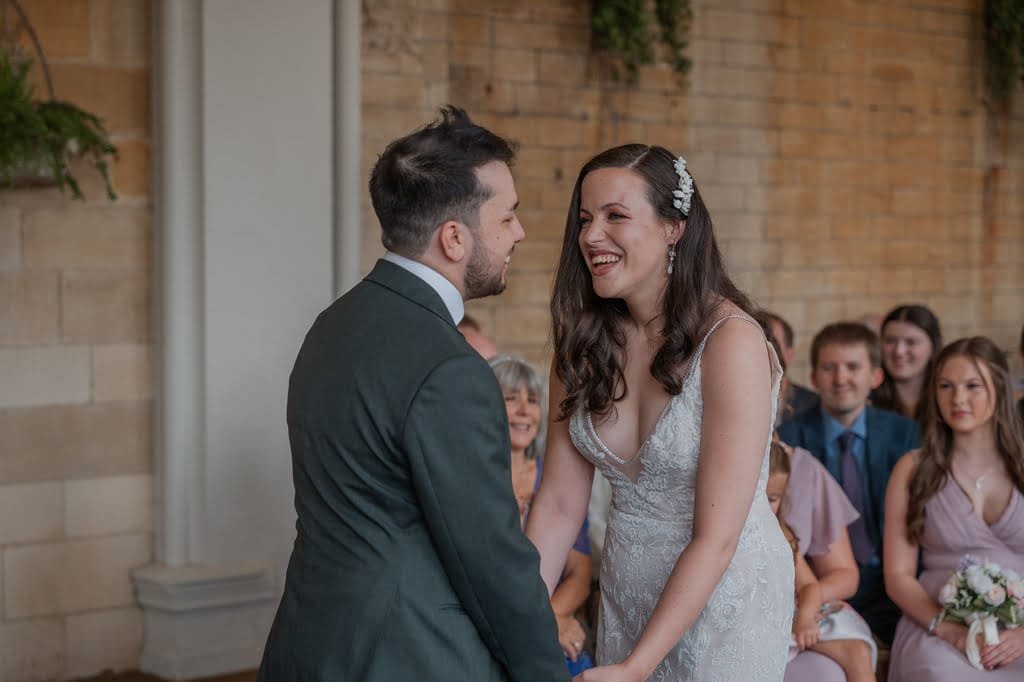 A couple smiling at each other while holding hands during a ceremony, captured by a Grittleton House Photographer. They are surrounded by seated guests in a room with a beige brick wall and greenery accents.