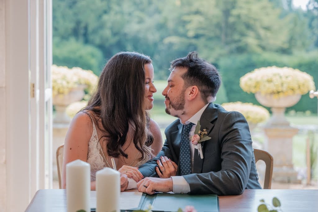 A couple sits closely together, holding hands and looking into each other's eyes in a bright room with large windows and a garden view. Candles are placed on the table in front of them, capturing a timeless moment as seen through the lens of a skilled Grittleton House photographer.