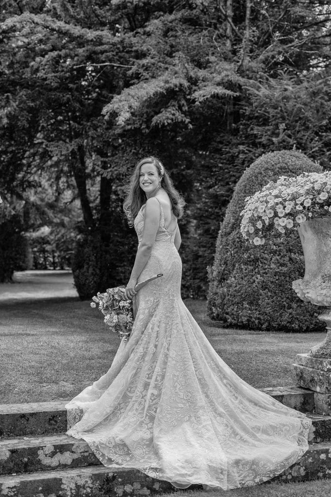 Black and white photo of a woman in a long, laced gown holding a bouquet of flowers, standing on stone steps surrounded by garden greenery. Captured with timeless elegance, it's the work of a talented Grittleton House photographer.