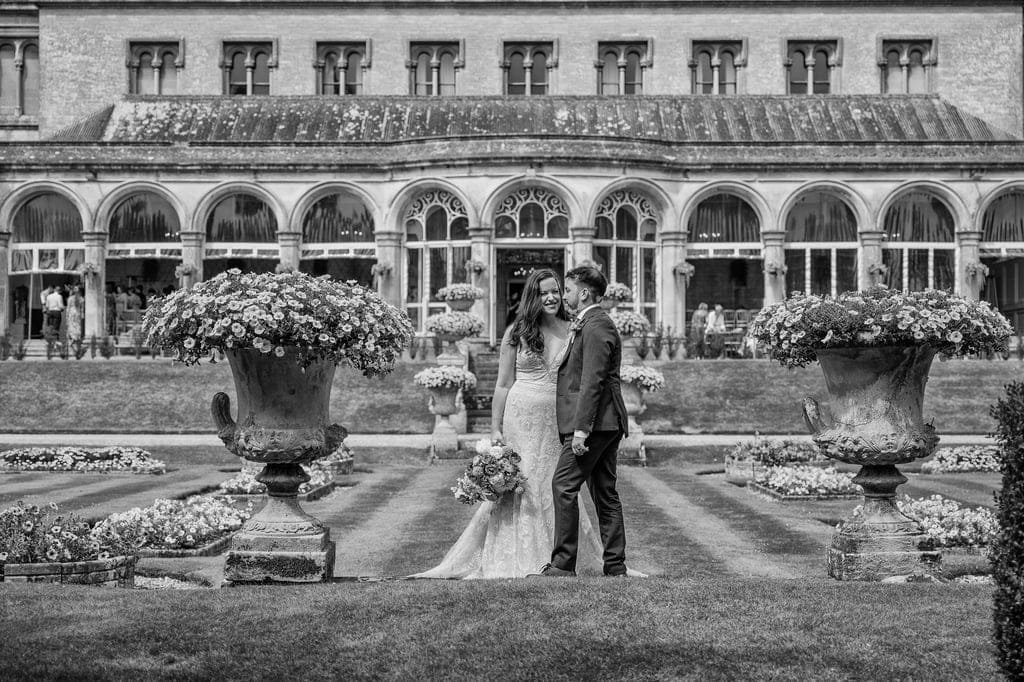 A bride and groom stand close together in front of the grand Grittleton House, a historic building with arched windows and manicured gardens in the background, holding a bouquet of flowers. Captured by their skilled photographer, the moment radiates timeless elegance.