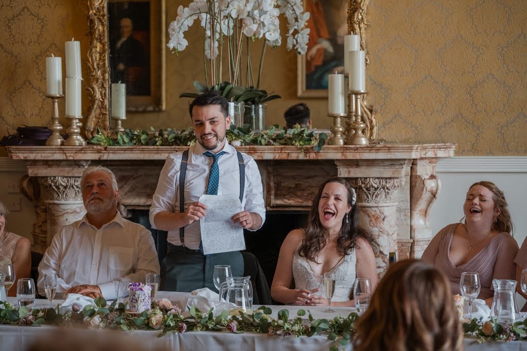 A man standing and speaking at a formal gathering, holding a piece of paper, with seated attendees around a table, including two women and an older man; all are smiling or laughing as captured by the Grittleton House Photographer.