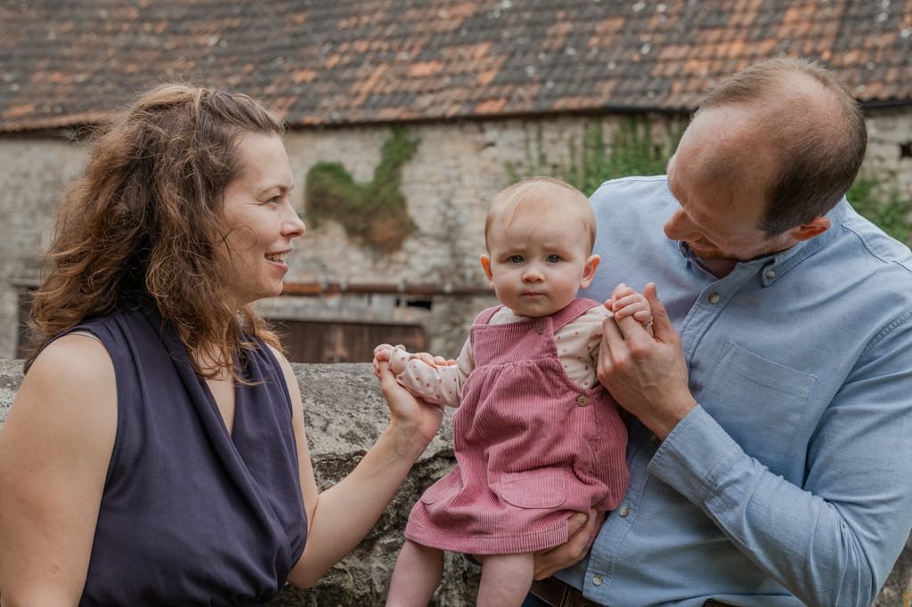 A woman and man hold a baby between them outdoors. The baby is dressed in a pink outfit while the adults smile at each other. Captured by a Frome portrait and wedding photographer, a rustic building is visible in the background.