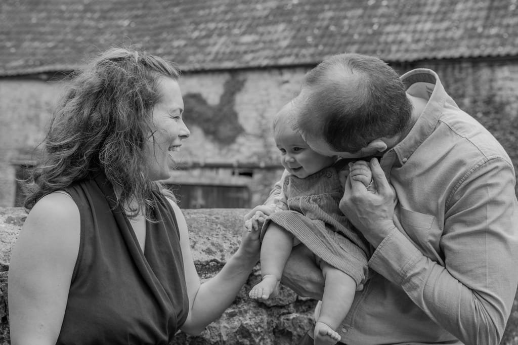 A man and woman stand next to each other, holding a baby. The man is playfully nuzzling the baby's face while the woman smiles at them. They are outdoors with a rustic building in the background, capturing a moment that could have been taken by a Frome portrait and wedding photographer.