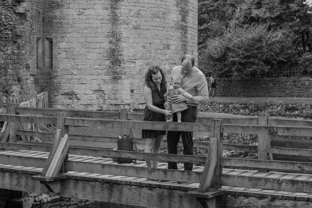 A man and woman hold a baby on a wooden bridge, with an old stone building and trees in the background. This enchanting scene, captured by a Frome Portrait photographer, exudes timeless charm.