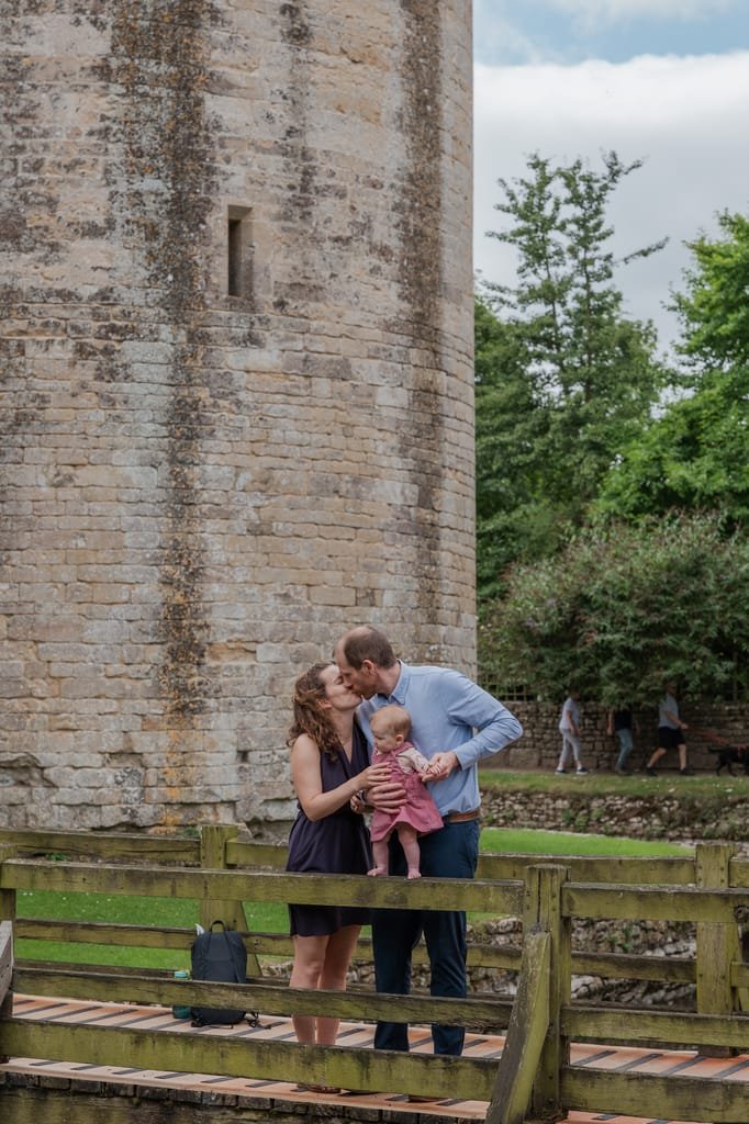 A couple stands on a wooden bridge in front of a stone tower, holding and kissing a baby. Trees and a few people are seen in the background, captured perfectly by a Frome wedding photographer.