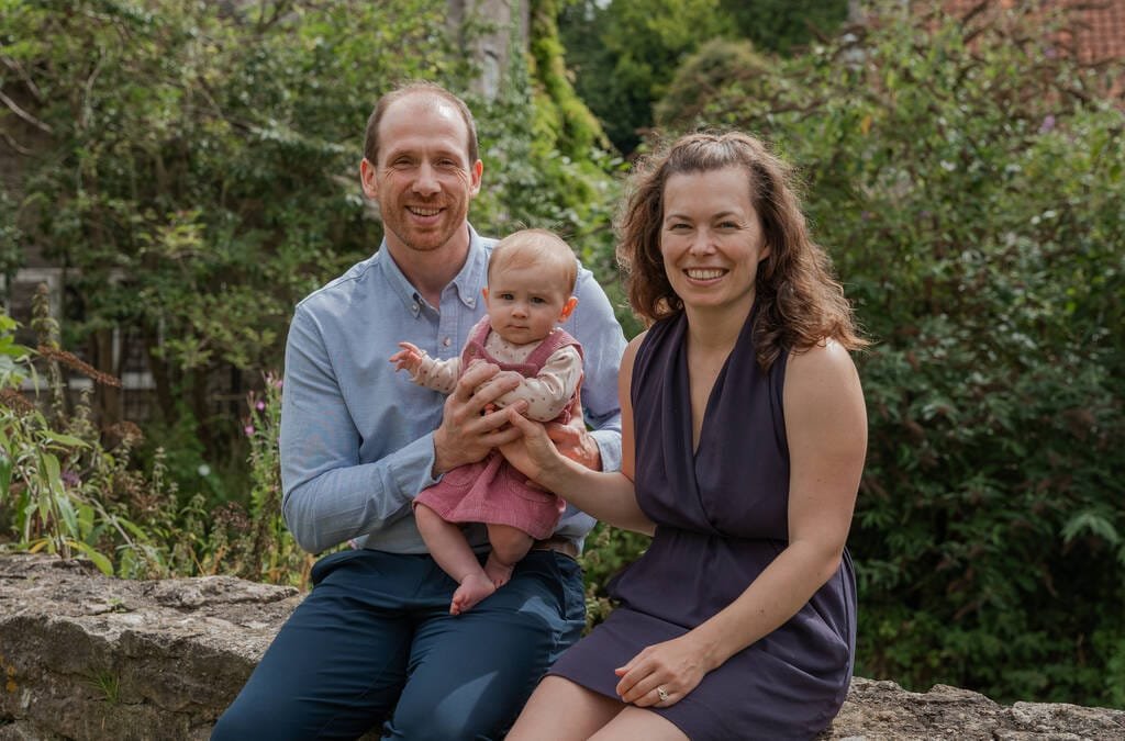 A man and woman sit outside holding a baby, smiling at the camera, with greenery and a house roof in the background—a perfect moment captured by a Frome portrait photographer.