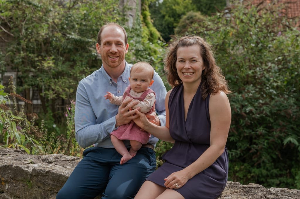 A man and woman sit outside holding a baby, smiling at the camera, with greenery and a house roof in the background—a perfect moment captured by a Frome portrait photographer.