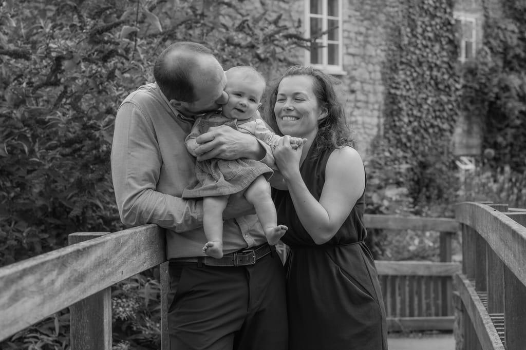 A man and a woman smile while holding a baby on a wooden bridge, with greenery and a stone building in the background, beautifully captured by Frome Portrait and Wedding photographer.