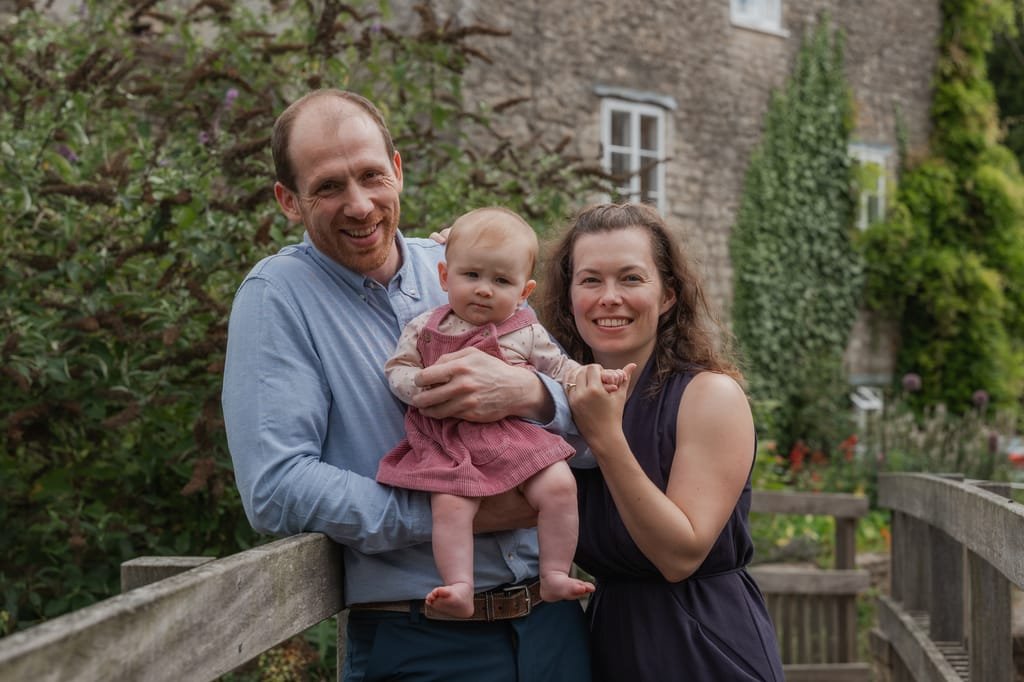 A man and woman stand outdoors, smiling at the camera, while holding a baby in a pink outfit. On a wooden walkway with lush greenery and a stone building in the background, this charming scene is captured by a talented Frome portrait photographer.