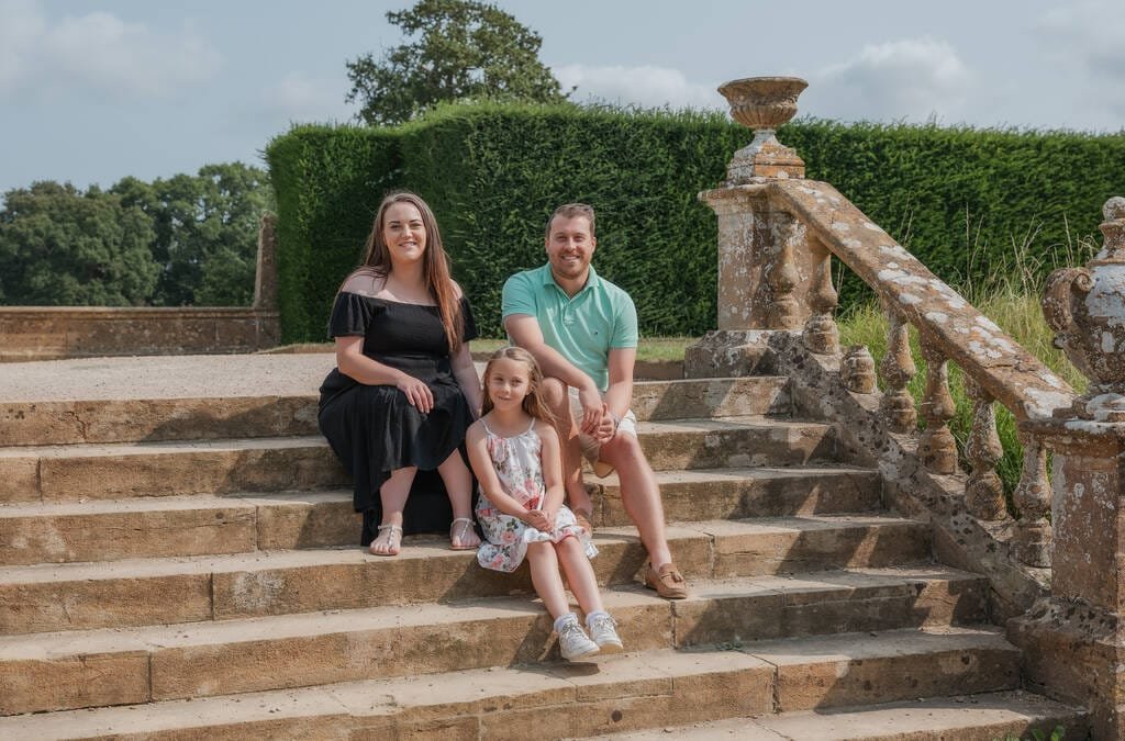 A woman, a man, and a child sit on stone steps outdoors at Montacute House. The woman and man are seated on either side of the child, surrounded by greenery and a stone railing in the background. This picturesque scene captures the essence of a National Trust family photoshoot.