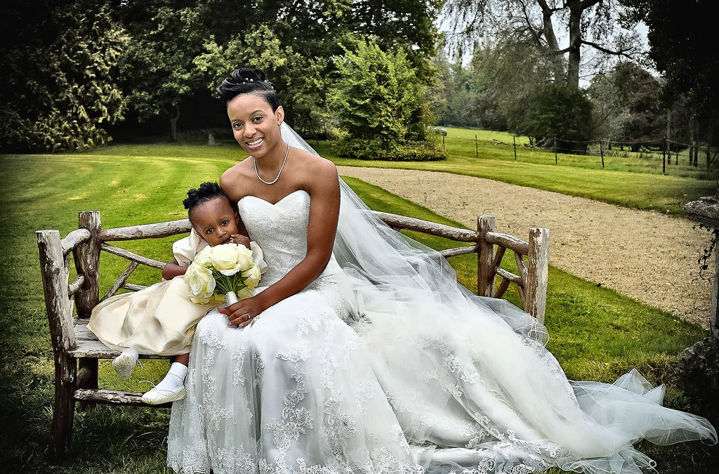 A woman in a wedding dress and a little girl sitting on a bench at Orchardleigh Wedding Photography.