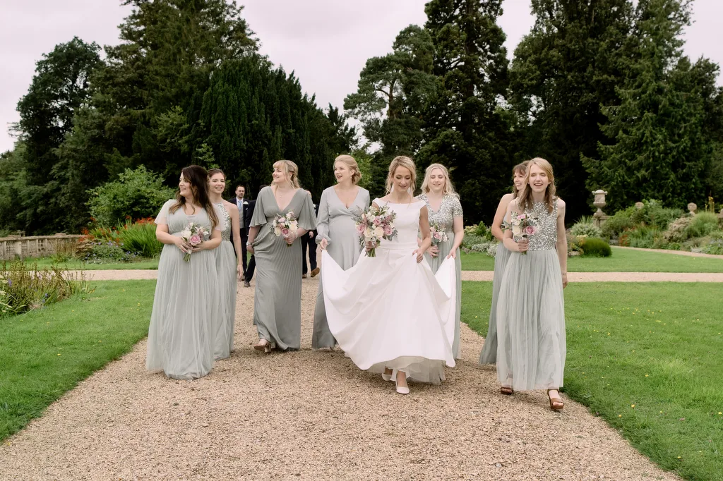 A bride and her bridesmaids gracefully walk through a captivating garden at Castle Combe Manor Wedding.