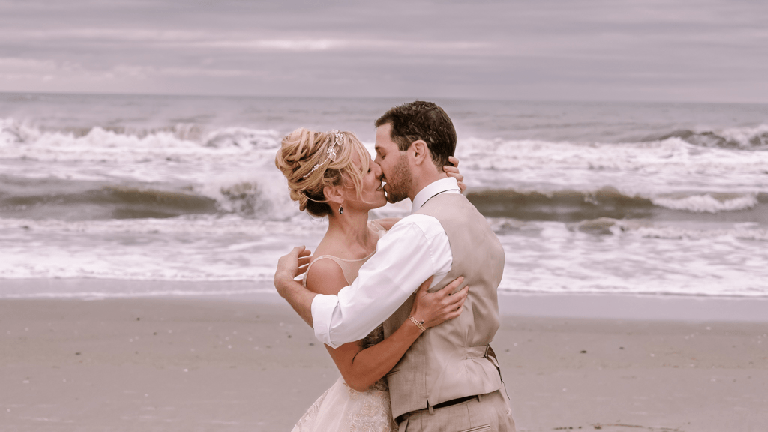 a bride and groom kissing on the beach.