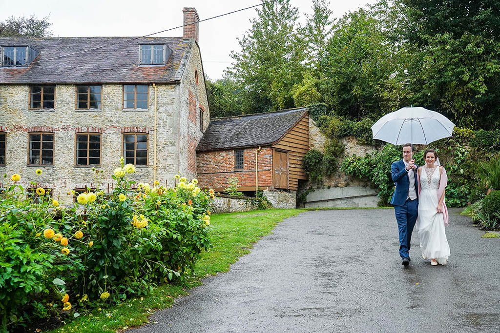 a bride and groom walking down a path holding an umbrella.