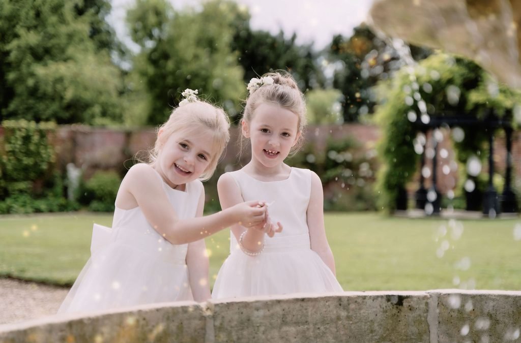 two little girls in white dresses standing next to a fountain.