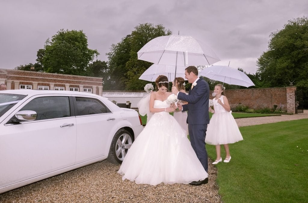 a bride and groom standing in front of a white car whist it rains