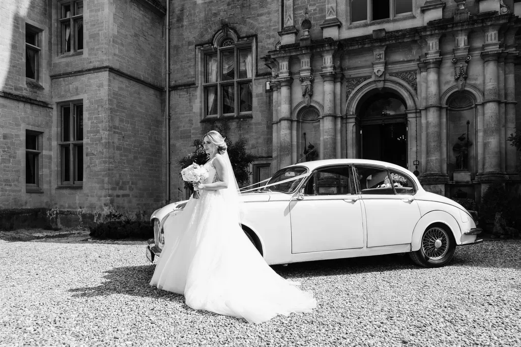 Orchardleigh `House:Cameras by Fuji:a bride standing next to a vintage car in front of a castle.