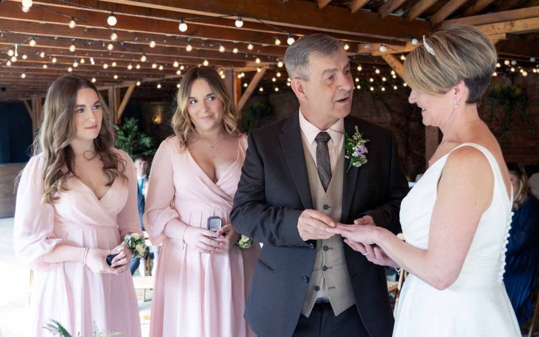 A man places a ring on a woman's finger during an Elmhay Park Weddings ceremony, while two women in pink dresses watch beside them under glowing string lights.