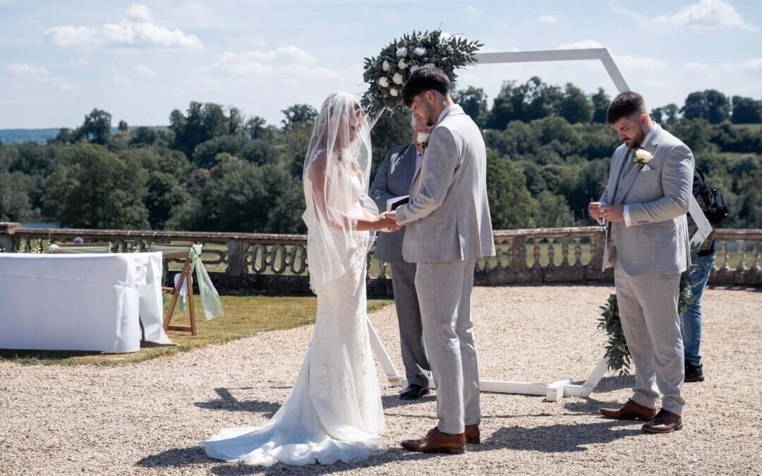 A bride and groom stand facing each other during an Orchardleigh House Weddings ceremony, with an officiant and groomsman nearby, under a beautifully decorated arch.