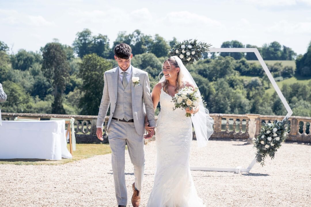 A bride and groom walk hand in hand outdoors at Orchardleigh House Weddings, smiling. The bride holds a bouquet; a floral arch and scenic greenery are visible in the background.