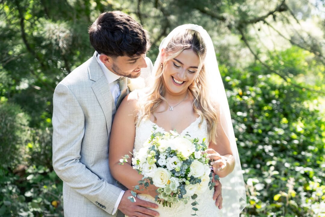 Bride and groom standing outdoors, smiling. The bride is holding a bouquet of white flowers, and the groom is leaning close behind her. Bright greenery in the background.