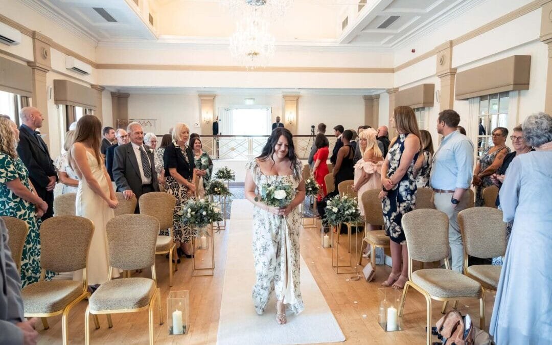 A woman in a floral dress walks down an aisle holding a bouquet, while guests seated on both sides watch in a brightly lit room with chandeliers at a stunning Bath Spa Hotel Weddings venue.
