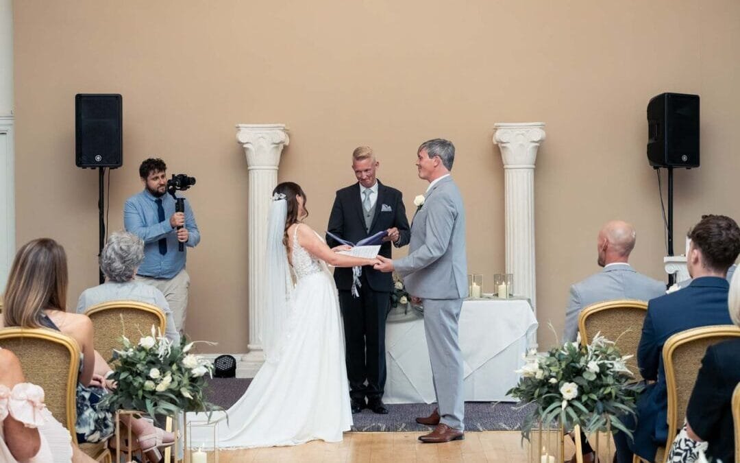 A bride and groom stand facing each other holding hands during a Bath Spa Hotel Weddings ceremony indoors, with guests seated and a photographer capturing the moment.