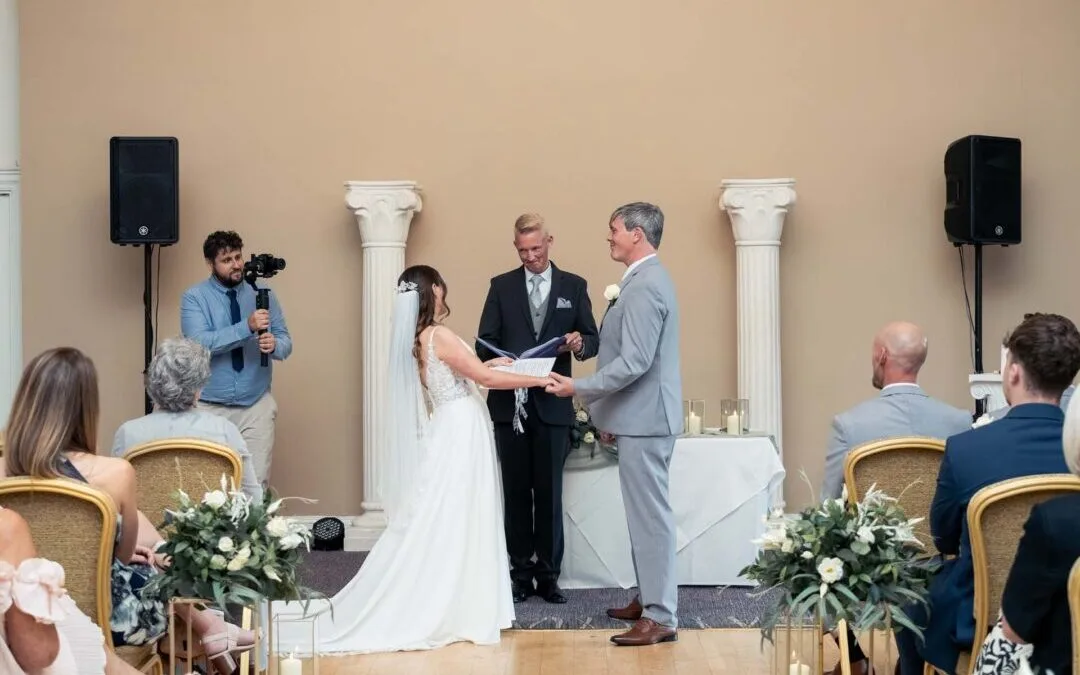 A bride and groom stand facing each other holding hands during a Bath Spa Hotel Weddings ceremony indoors, with guests seated and a photographer capturing the moment.