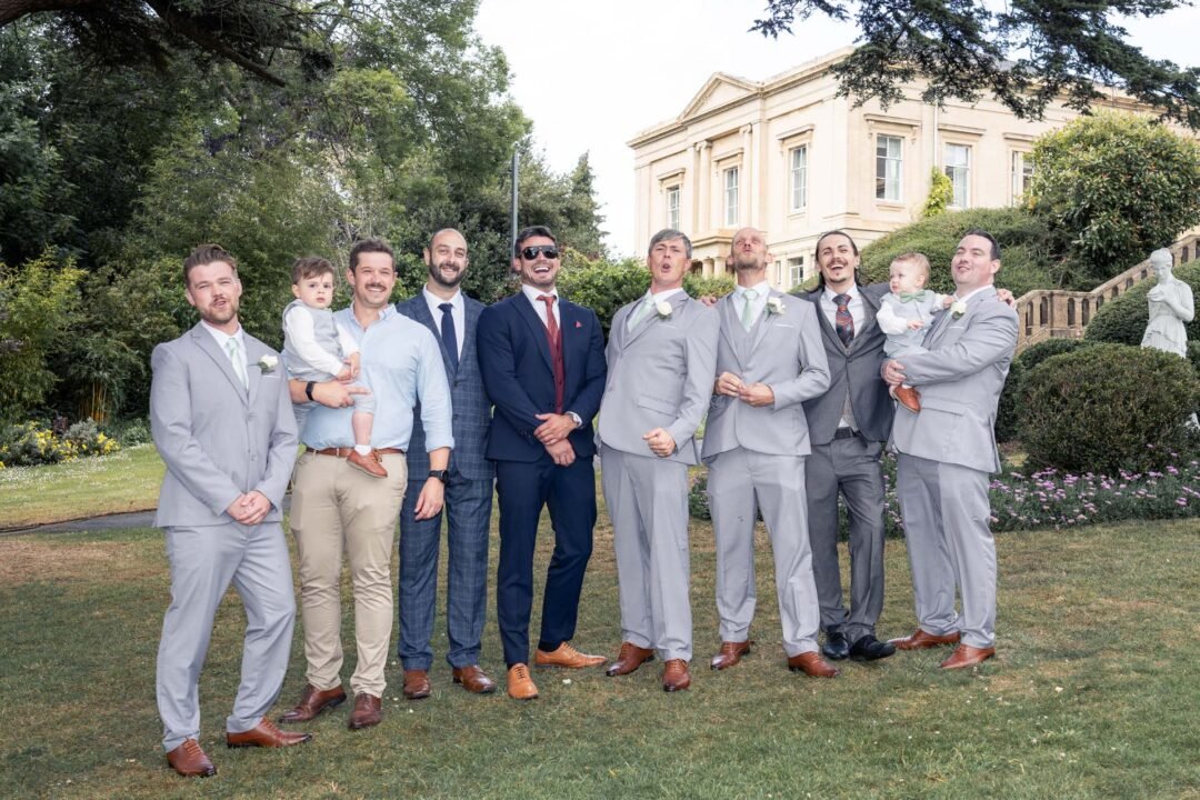 20250618L&E0906 A group of men and two young children pose for a formal photo on a lawn in front of a large building, dressed in suits and ties—capturing the elegance often seen at Bath Spa Hotel Weddings.