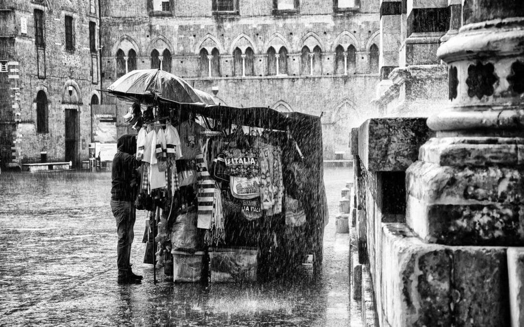A street vendor stands under an umbrella by a stall selling souvenirs in heavy rain near a historic stone building, captured with the timeless contrast of black & white film photography.