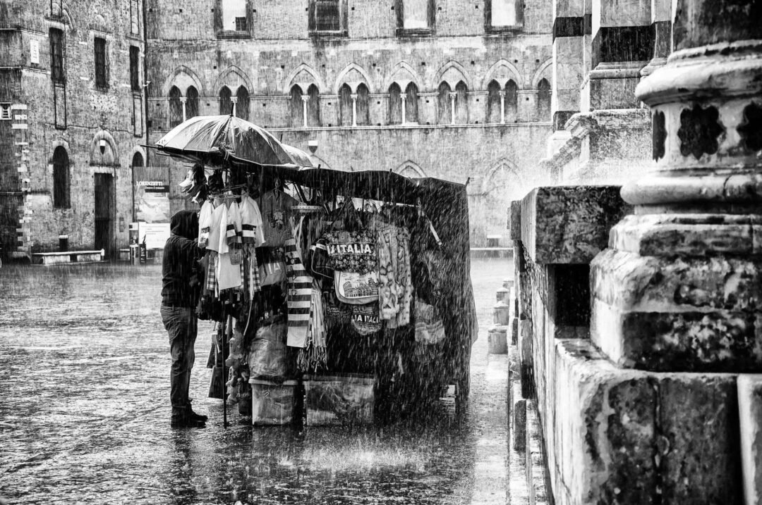 A street vendor stands under an umbrella by a stall selling souvenirs in heavy rain near a historic stone building, captured with the timeless contrast of black & white film photography.