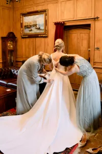 Two bridesmaids helping each other put on their wedding dresses at The Manor in Castle Combe, captured by a talented Wedding Photographer.