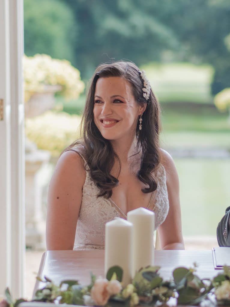 A woman with long brown hair, wearing a white dress and floral hair accessory, sits at a table adorned with white candles and greenery, smiling. She is outdoors with a blurred garden background, captured beautifully by a Grittleton House photographer.