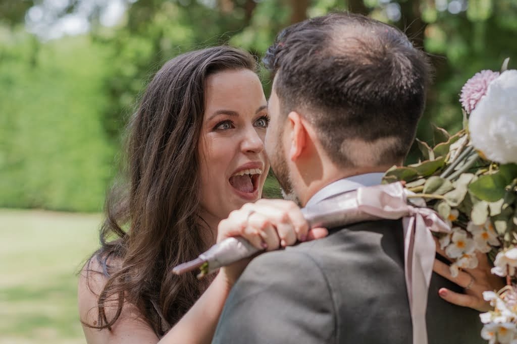 A woman with long hair excitedly smiles at a man in a suit holding a bouquet of flowers. They are outdoors, surrounded by lush greenery, caught beautifully by the Grittleton House photographer.